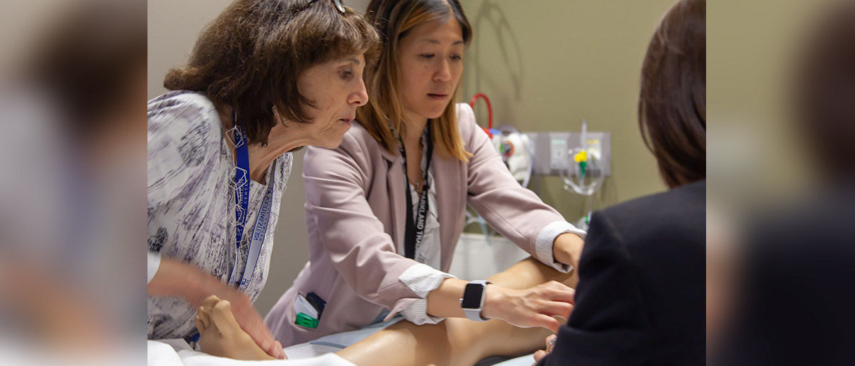 Three women gathered around a human simulation dummy, one woman is demonstrating something with her hands