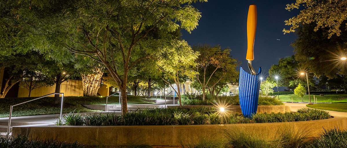 Scultpure of a trowel with a blue blade and sand colored handle, positioned amongst greenery at night