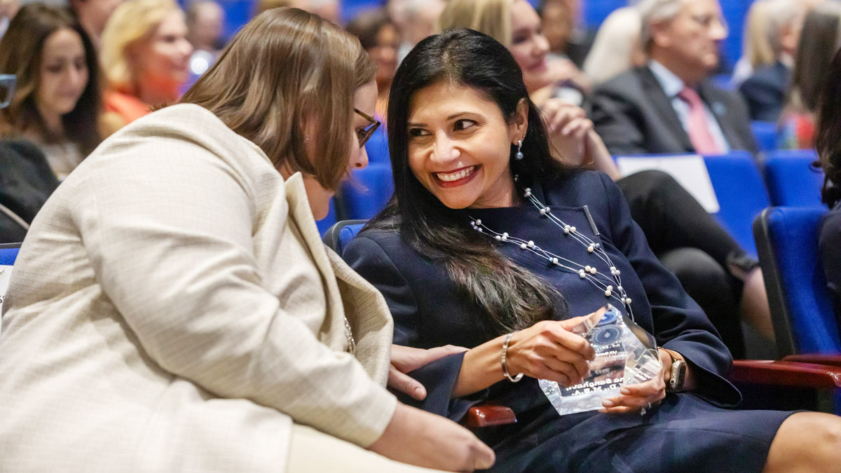 Two women seated in auditorium, chatting.