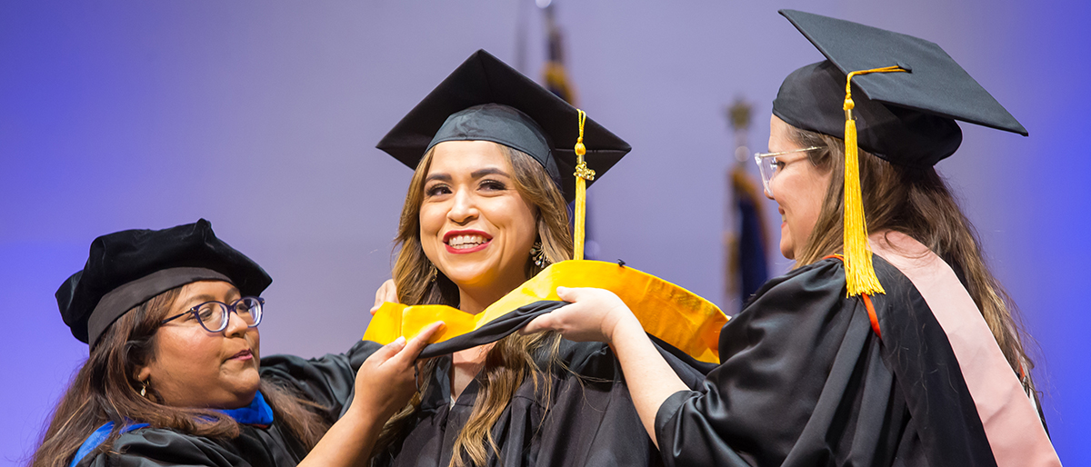 Three women dressed in graduation cap and gown. Two of the woman are placing a yellow sash on the woman in the center.