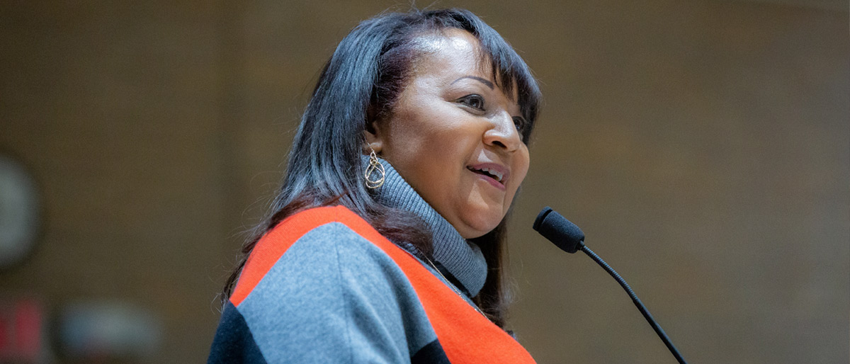 Woman wearing an orange and slate sweater, speaking into a microphone