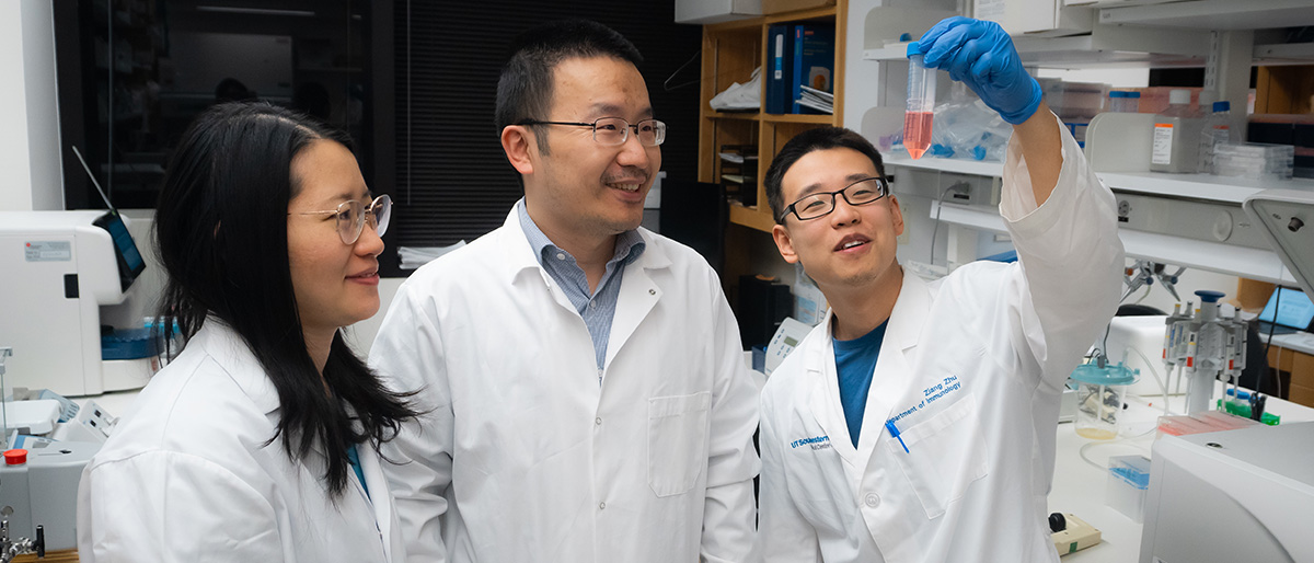 Smiling Dr.s Yao, Wu and Zhu wearing lab coats in lab looking at test tube held up by Dr. Zhu.