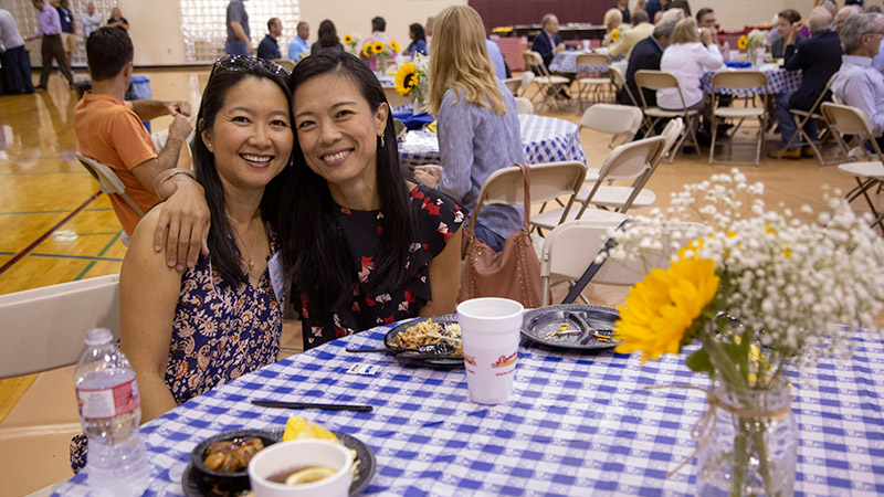 Two women seated and smiling for the camera, one with her arm around the other.