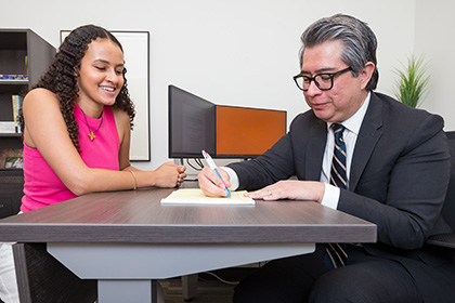 Smiling woman with wavy brown hair wearing a pink shirt watching a man with graying hair and glasses wearing and a dark suit write on a note pad.