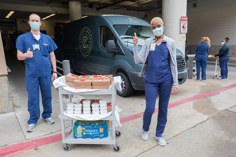 Two people in scrubs giving thumbs up next to cart of food