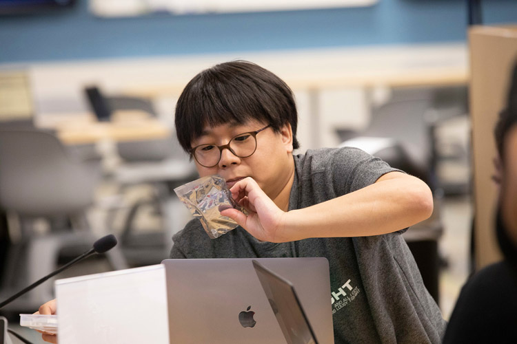 Person with glasses, black hair holding an item while standing over their computer