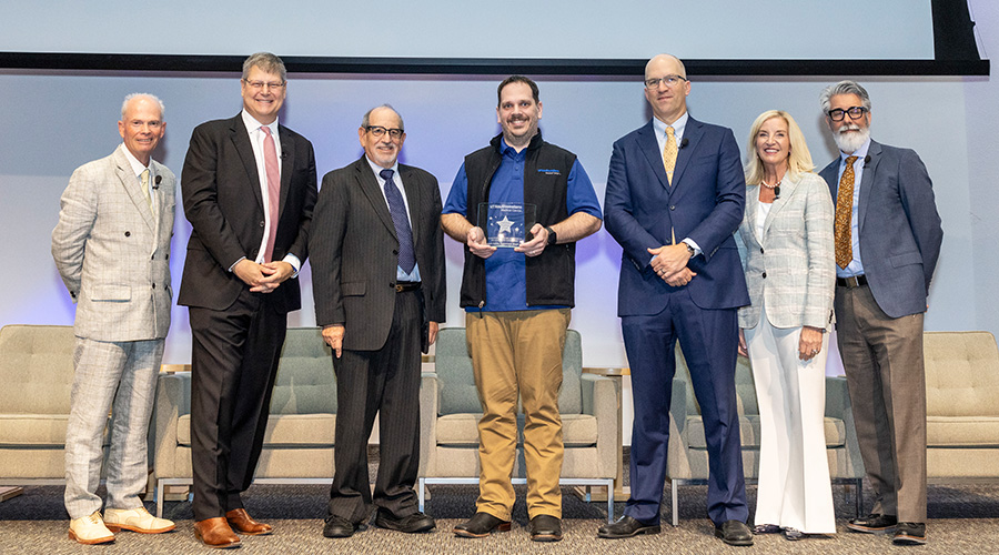 6 speakers stand with award winner in center holding plaque-from left, man in tan suit, tall man in black suit pink tie, shorter man in black suit, man in blue shirt holding award, bald man in blue suit, blonde woman in cream suit, bearded man in grey and tan suit