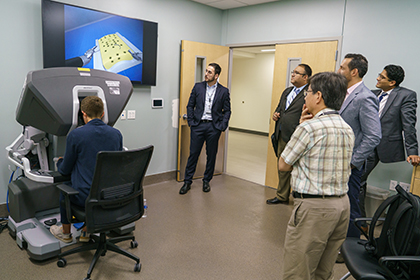 A seated person uses a simulation machine to make a suture and a group of men watch the the process on a large monitor.