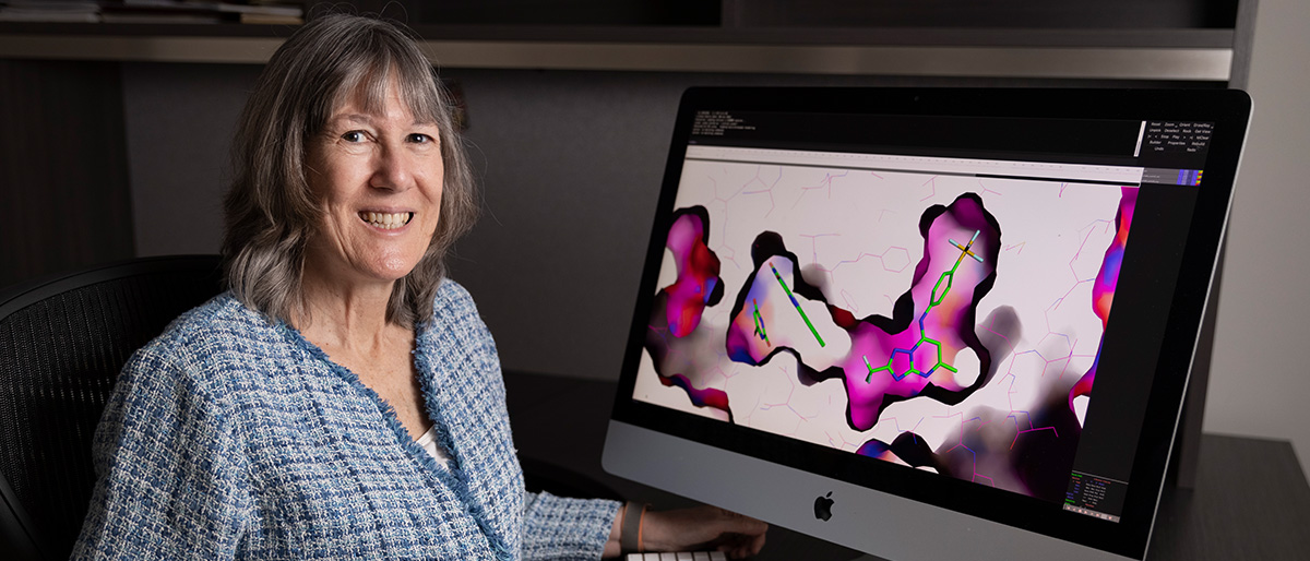 Smiling woman with salt-and-pepper hair wearing a blue and gray plaid shirt sitting in front of a large computer monitor with molecular diagrams in black, pink and green on the screen.