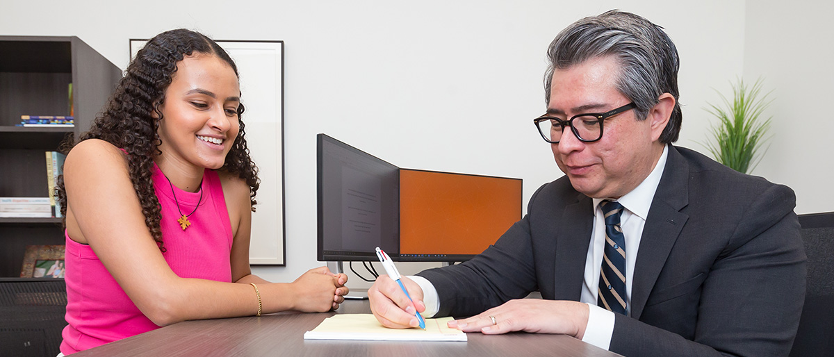 Smiling woman with wavy brown hair wearing a pink shirt watching a man with graying hair and glasses wearing and a dark suit write on a note pad.