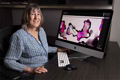 Smiling woman with salt-and-pepper hair wearing a blue and gray plaid shirt sitting in front of a large computer monitor with molecular diagrams in black, pink and green on the screen.