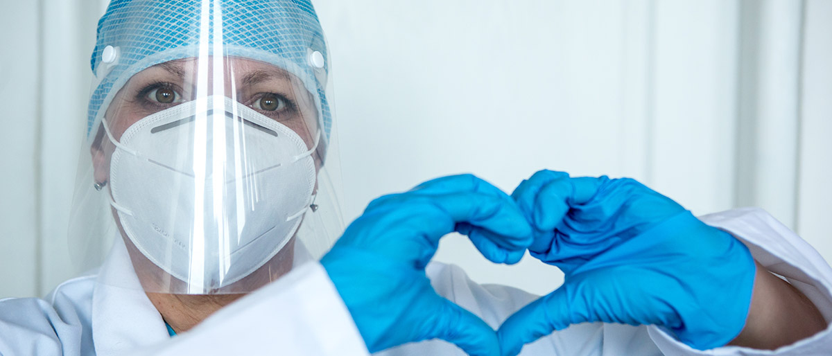Person in mask and white coat making heart shape with hands