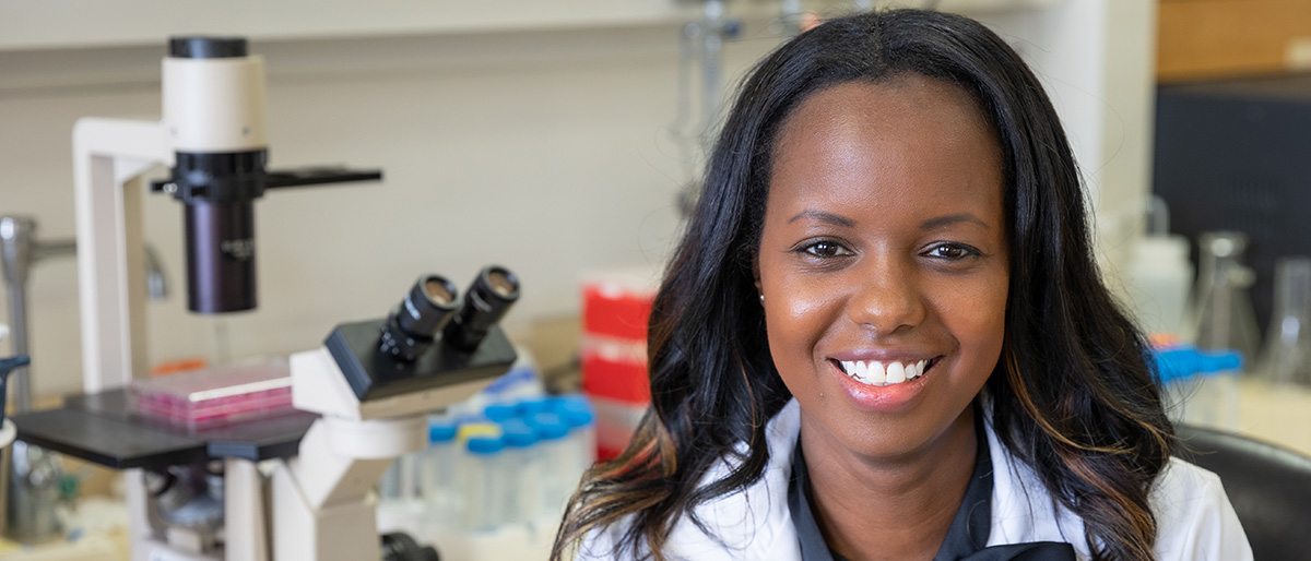 Smiling woman with long dark hair, wearing a lab coat. Microscope is on the desk in the background.