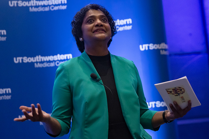 female speaker on stage, dark curly hair, wearing green jacket and black shirt