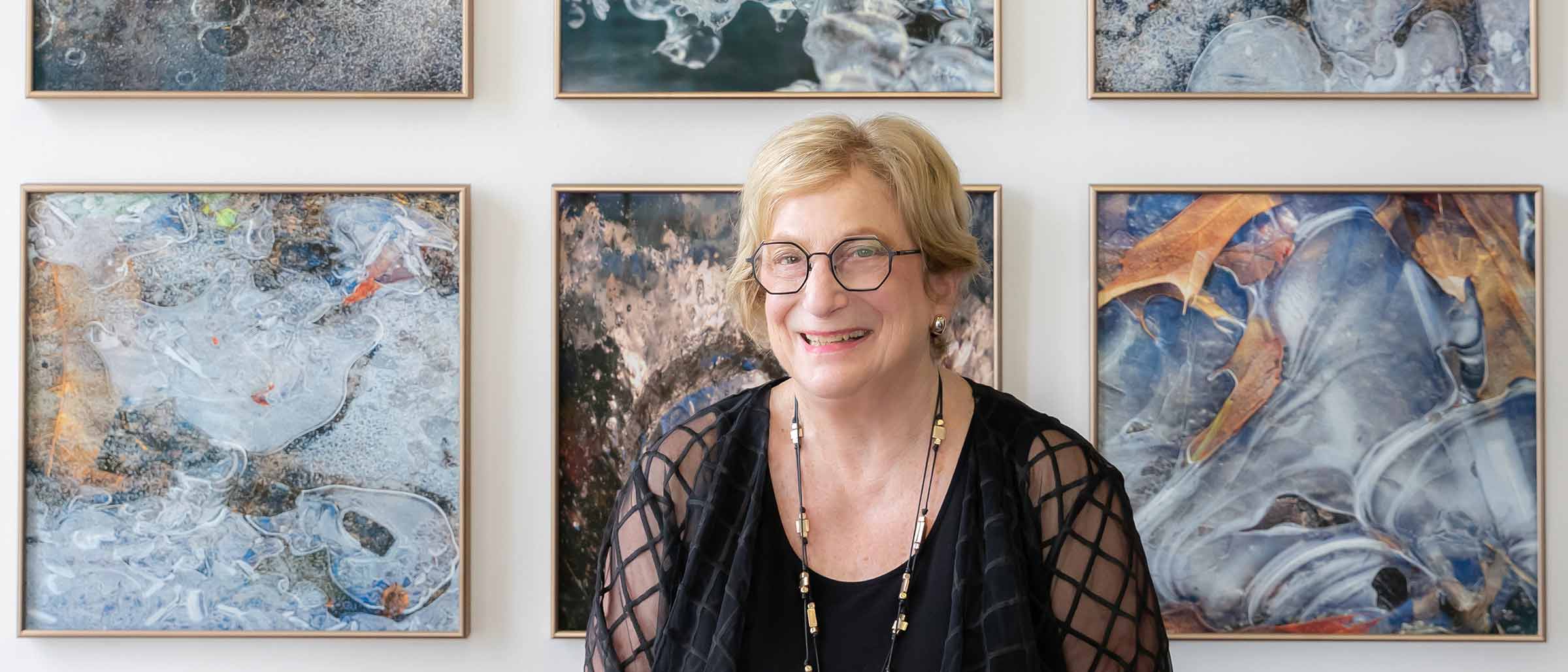 Woman with short blonde hair, black dress, seated in front of art