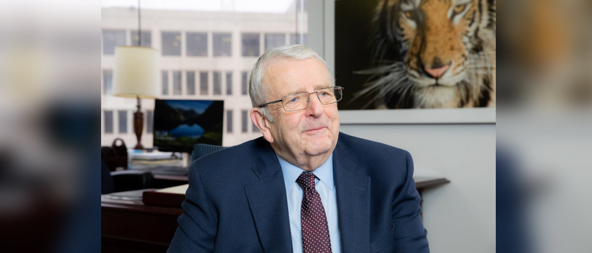 Man with grey hair, glasses, in a dark blue suit sitting in an office