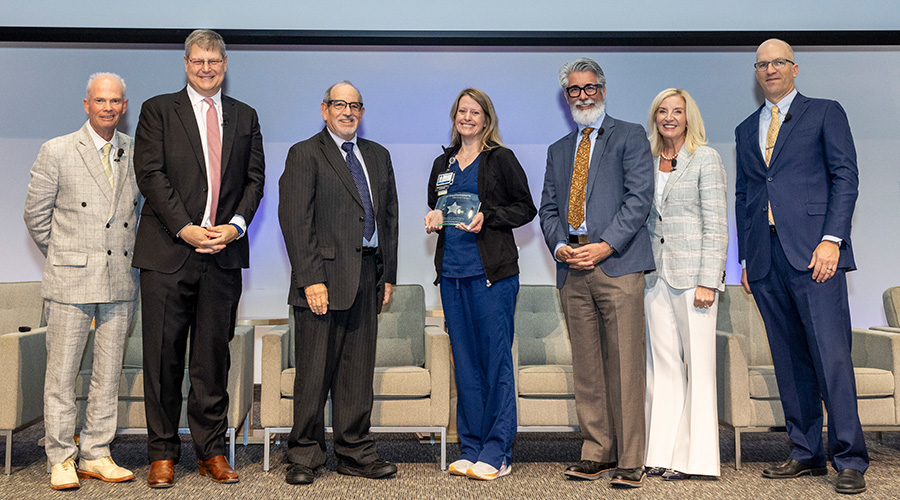 6 speakers stand with award winner in center holding plaque-from left, man in tan suit, tall man in black suit pink tie, shorter man in black suit, woman holding award in blue scrubs, bearded man in grey and tan suit, blonde woman in cream suit, bald man in blue suit