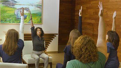 A group of 5 women sitting on chairs, following a leader doing chair yoga.
