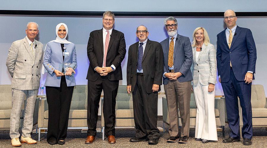 7 speakers standing on stage- man in tan suit, woman in white burka w/blue blazer, tall man in black suit pink tie, short man in black suit, grey bearded man in gray and tan suit, blonde woman in cream suit, balding man with glasses in blue suit
