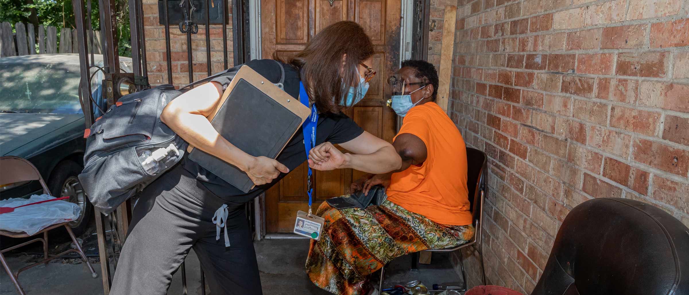 Woman in scrubs and mask giving an elbow bump greeting to a woman in mask, face shield seated on her front porch