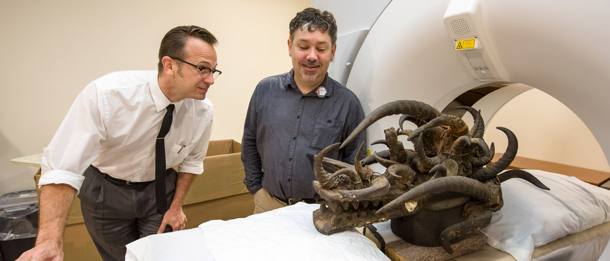 Two people standing next to a CT scanner with an ornate animal head mask on it