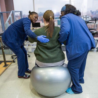 Two women in scrubs assist a patient sitting on a yoga ball