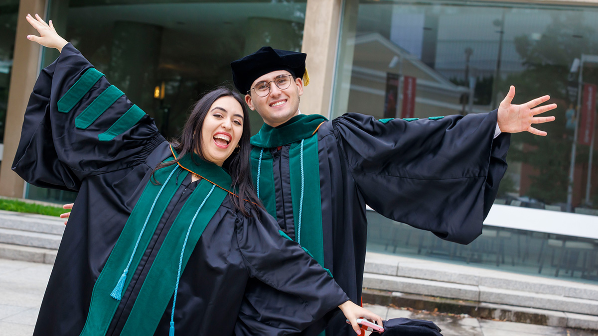 Two student wearing black and green robes, wave their arms as if spreading their wings.