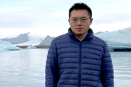 Dark-haired man wearing glasses and a blue puffer jacket standing in front of glacier and ice flow.