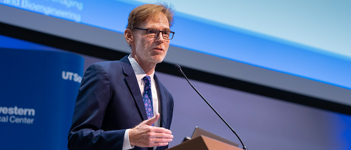 Man with fair hair, wearing glasses and a dark suit, lecturing from a podium.