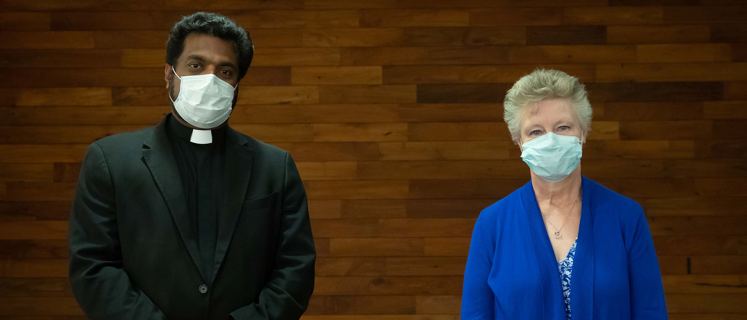 Man in black jacket and priest's collar and woman in blue, wearing masks and standing apart, in wood paneled room