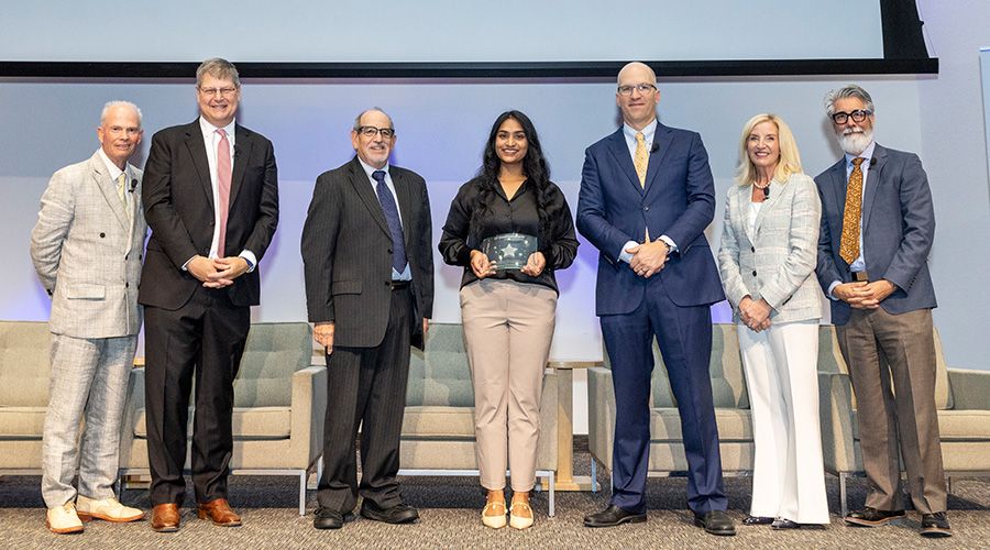 award winner in center holds plaque - from left, man in tan suit, tall man in black suit pink tie, shorter man in black suit, woman with long dark hair holds award, bald man in blue suit, blonde woman in cream suit, bearded man in grey and tan suit