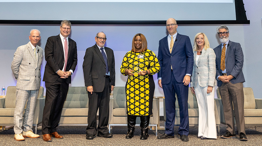 6 speakers stand with award winner in center holding plasuique-from left, man in tan suit, tall man in black suit pink tie, shorter man in black suit, woman in yellow and black dress holding award, bald man in blue suit, blonde woman in cream suit, bearded man in grey and tan suit