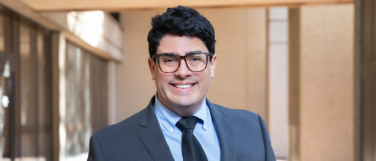 Man in grey suit, navy tie, with glasses and black hair