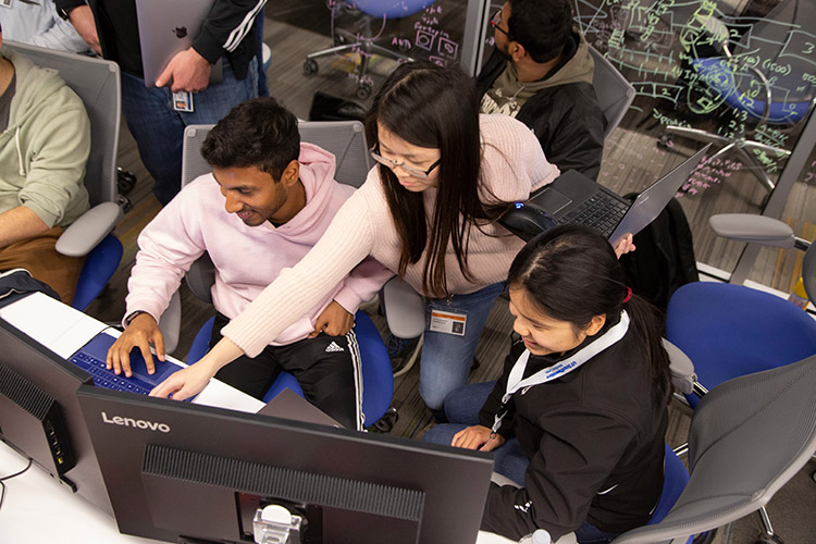 Three people gathered around computers, one standing and holding hers as she points at another's screen