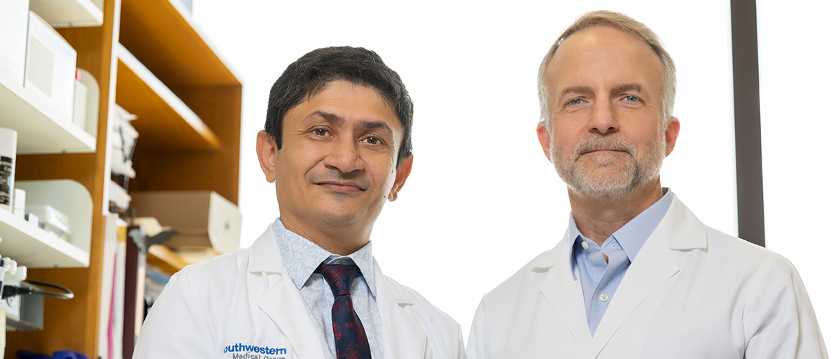 Dr Patel, left, with dark hair, wearing a lab coat and dark tie. Dr. Buszczak, right, with gray hair and beard, wearing a lab coat. They are standing in a lab.