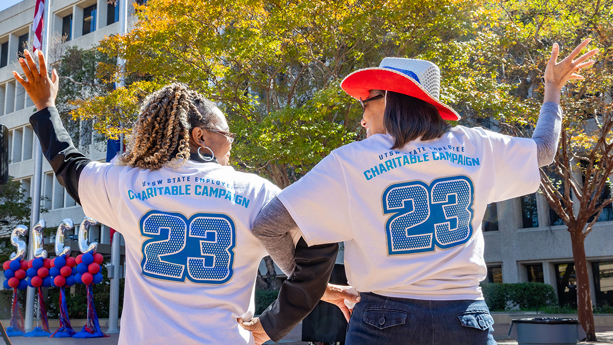 Backs of two women standing outside wearing matching jerseys, that say Charitable Campaign 23.