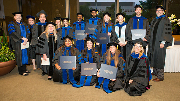 Group of graduates wearing black robes and morter boards, and blue sashes, holding their diplomas or programs.