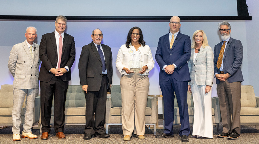 6 speakers stand with award winner in center holding glass plaque- from left, man in tan suit, tall man in blue suit pink tie, shorter man in black suit, dark haired woman in white and tan holding award, tall man in blue suit, blond woman in cream suit, bearded man in grey and tan suit