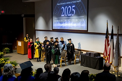 Speakers on stage at the Peter O'Donnell Jr. School of Public Health 2025, Commencement Exercises face the flags with right hands over their hearts.