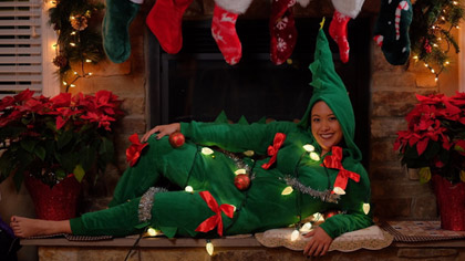 Woman dressed in green onesie covered in bows and lights reclining on a fireplace