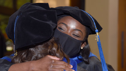 Woman in graduation cap and gown and mask hugging another