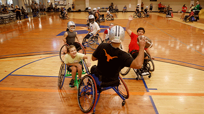 Peope iin wheelchairs ranging in age from children to adults play football in a gym. Some are wearing Dallas Cowboy helmits.