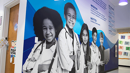 Wall mural at Biomedical prep school. 5 students in the forground dressed in medical garb, wearing stethoscopes, one holds a computer. Behind them, part of the mural's message is visable