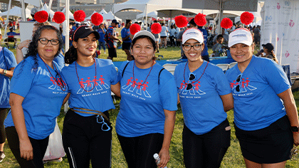 Five dark-haired women wearing matching blue t-shirts. The three on the right are wearing white UT Southwesterrn caps, second from the left is wearing a black cap. All 5 are wearing headbands with red pom-poms.