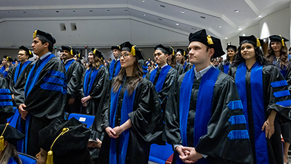 audience standing together, all graduates wearing black and blue cap and gown