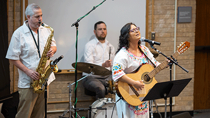 Woman with long dark hair and glasses wearing a colorfully emboidured dress, plays guitar and sings. In the background one man plays the saxaphone and another plays the drums.