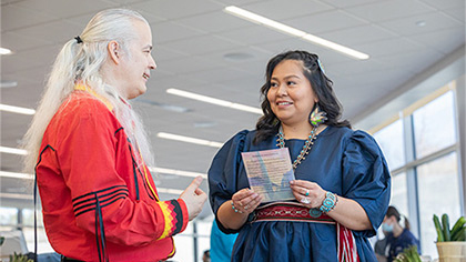 Man with long gray hair wearing a red jacket talking to a woman with long dark hair wearing a blue dress. She is also wearing colorful beaded earrings and a necklace, rings and bracelets made of silver and turquoise. The woman is holding what could be a program from the event.