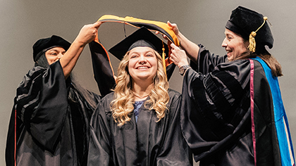 young woman wearing cap and gown receives gold sash from 2 other women during graduation ceremony