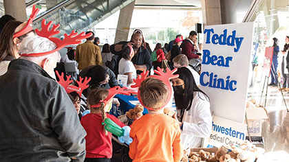 Adults and children wearing red foam reindeer antlers, watch young woman with long dark hear wearing PPE work on a teddy bear at a table with a sign that says, 
