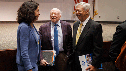 A woman with shoulder-length dark, curly hair wearing a blue suit, chatting with two older white-haired men wearing suits and ties. One man is holding notebooks and the other is holding the event program.
